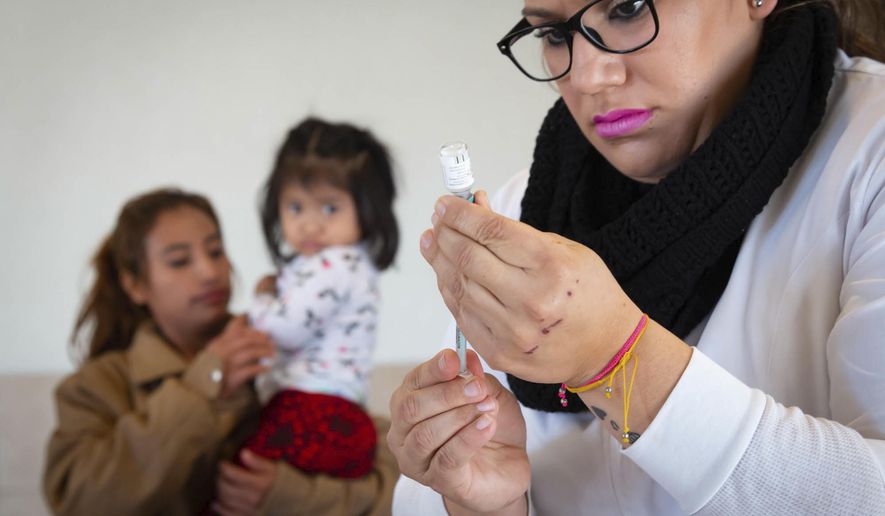 In this Jan. 28, 2020, photo provided by Cronkite News, Arizona State University, Andrea Ramirez, 2, of Chiapas, Mexico, watches Daniela Gastelum as she prepares a vaccine at Casa del Migrante in San Luis Rio Colorado, Mexico. One Hundred Angels, a Phoenix organization that provides medical care and other services to migrants, helped coordinate the one-day vaccination clinic at Casa del Migrante, working with the Mexican Red Cross. (Delia Johnson/Cronkite News, Arizona State University via AP)