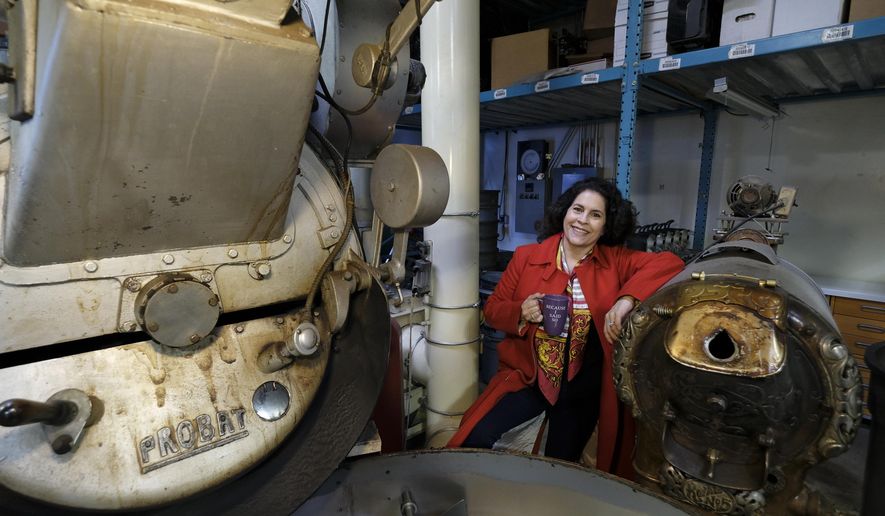 In this photo taken Wednesday, March 11, 2020, Renee Brown, vice-president of Wild Card Roasters, poses between a pair of coffee roasters at her roastery and cafe in San Rafael, Calif. As the coronavirus spreads, many small business owners are rethinking their strategies and adjusting the way they work. (AP Photo/Eric Risberg)
