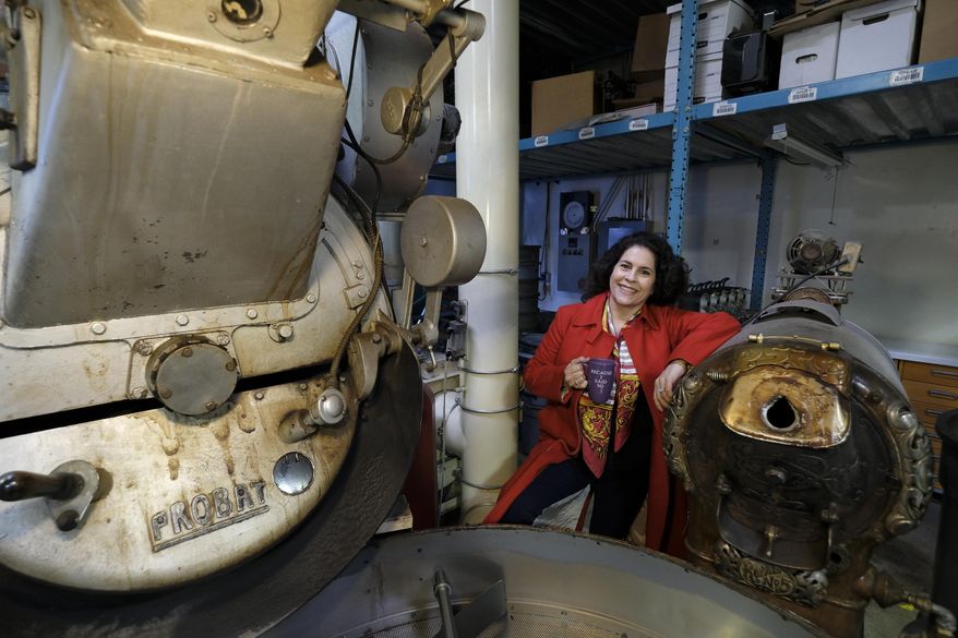 In this photo taken Wednesday, March 11, 2020, Renee Brown, vice-president of Wild Card Roasters, poses between a pair of coffee roasters at her roastery and cafe in San Rafael, Calif. As the coronavirus spreads, many small business owners are rethinking their strategies and adjusting the way they work. (AP Photo/Eric Risberg)
