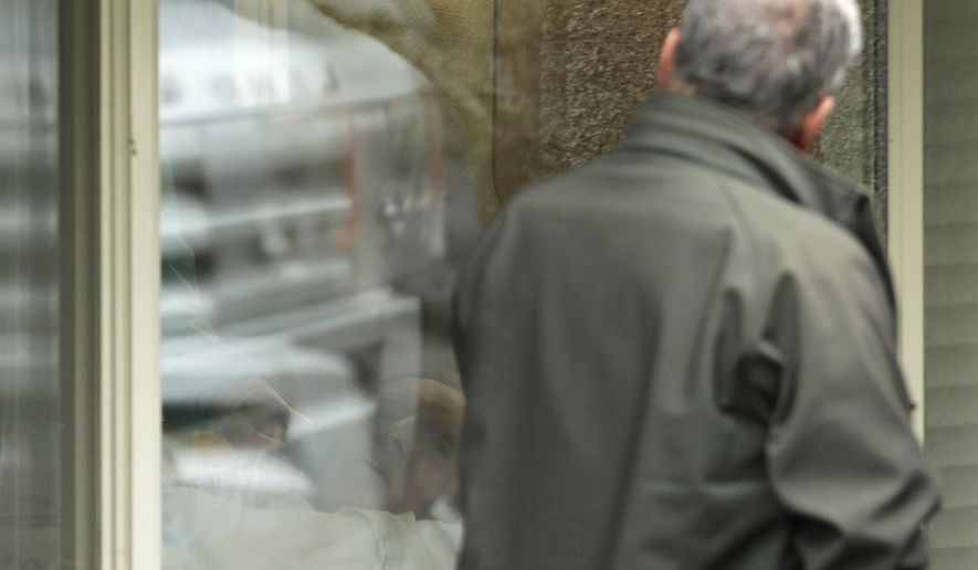 In this March 12, 2020, photo, Steve Sedlacek, right looks at his father, Chuck Sedlacek, back left, through reflections in the window of his room at Life Care Center in Kirkland, Wash., near Seattle. The facility has been at the center of the coronavirus outbreak in the state, and Sedlacek's brother Scott said that he and his siblings have barely spoken to their father inside the center, who in addition to testing positive for the coronavirus, has blindness, neuropathy, and has difficulty using a phone, saying he is more of an "inmate" than a patient. Residents of assisted living facilities and their loved ones are facing a grim situation as the coronavirus spreads across the country, placing elderly people especially at risk. (AP Photo/Ted S. Warren)
