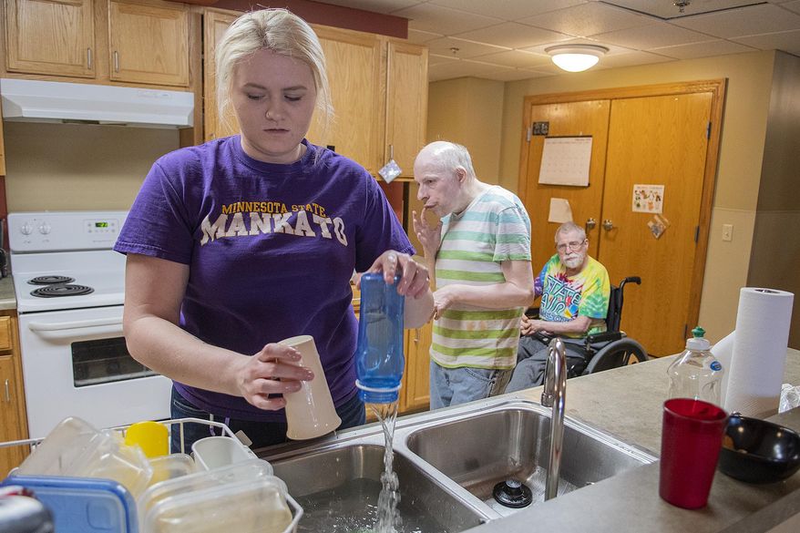 In this Feb. 27, 2020 photo, Wylie Rollings does the dishes in the kitchen in a community room at the Harry Meyering Center in Mankato, Minn., as residents Michael Dreyer and Richard Anderson, right, mull about behind her. (Jackson Forderer/The Free Press via AP)