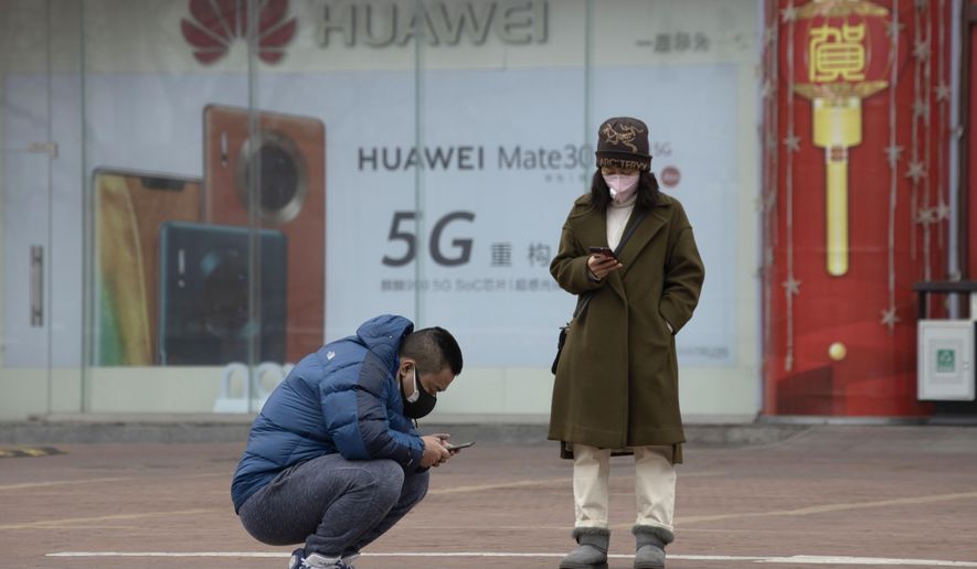 In this Sunday, March 8, 2020, photo, residents wearing masks look at their smartphones near an advertisement for 5G smartphones from Chinese tech giant Huawei in Beijing. Factories in China, struggling to reopen after the coronavirus shut down the economy, face a new threat from U.S. anti-disease controls that might disrupt the flow of microchips and other components they need. For most people, the new coronavirus causes only mild or moderate symptoms. For some it can cause more severe illness. (AP Photo/Ng Han Guan)