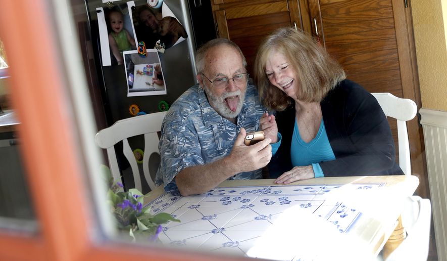 Seen through their kitchen window, Allan and Debbie Cameron contact their grandchildren via the internet Wednesday, March 25, 2020, in Chandler, Ariz. Debbie, 68, has asthma which makes her one of the people most at risk from the new coronavirus. The Cameron's now she see their children and grandchildren from the other side of a window or a phone. (AP Photo/Matt York)