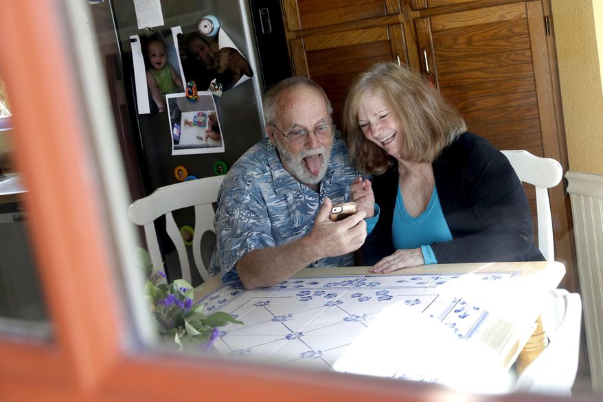 Seen through their kitchen window, Allan and Debbie Cameron contact their grandchildren via the internet Wednesday, March 25, 2020, in Chandler, Ariz. Debbie, 68, has asthma which makes her one of the people most at risk from the new coronavirus. The Cameron's now she see their children and grandchildren from the other side of a window or a phone. (AP Photo/Matt York)