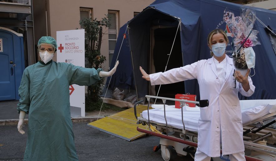 Monica Carfora, right, deputy head of the emergency of the Santo Spirito Hospital, which now has a separate emergency for possible covid patients, holds a chocolate Easter egg as she poses for a photograph with nurse Silvia Sforza, holding out their arms to indicating social distancing, outside the entrance to for covid emergency, in central Rome, Sunday, April, 12, 2020. The new coronavirus causes mild or moderate symptoms for most people, but for some, especially older adults and people with existing health problems, it can cause more severe illness or death. (AP Photo/Alessandra Tarantino)