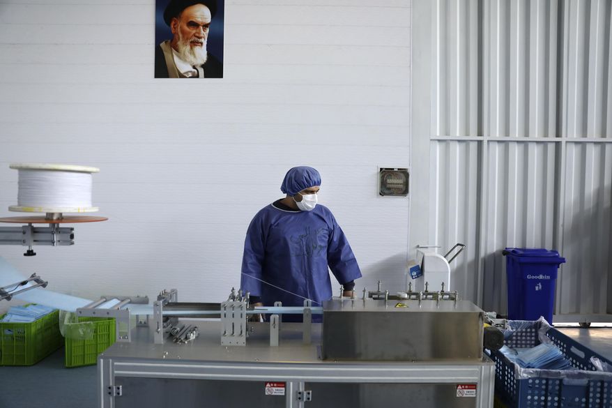 Under a portrait of the late Iranian revolutionary founder Ayatollah Khomeini, a worker wearing a protective suit controls a machine which makes face masks at a factory in Eshtehard, some 70 miles (114 kilometers) west of the capital Tehran, Iran, Tuesday, April 14, 2020. The factory is producing protective masks round-the-clock in a fight against the new coronavirus to meet the domestic demand as the country faces the worst outbreak in the Middle East. (AP Photo/Vahid Salemi)
