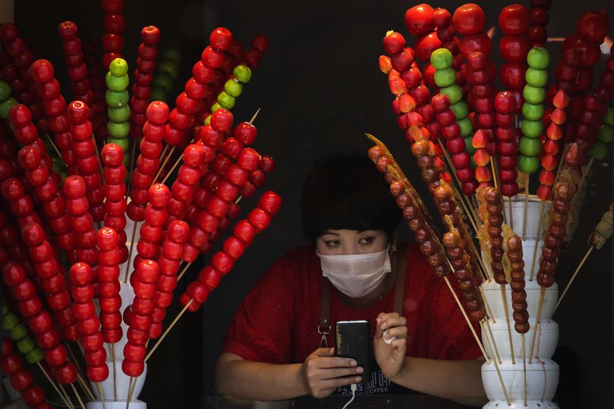 A vendor wearing a protective face mask looks out of her store selling sugar-coated haws on a stick in Beijing, Sunday, April 12, 2020. The new coronavirus causes mild or moderate symptoms for most people, but for some, especially older adults and people with existing health problems, it can cause more severe illness or death. (AP Photo/Andy Wong)