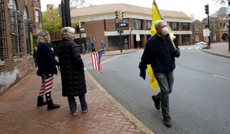 A man carries a flag as he walks by two protestors gathered at Church Circle, Monday, April 20, 2020, in Annapolis, Md. Only a few people showed up to protest and call for the state government to reopen amidst the coronavirus outbreak. Over the weekend, many protestors riding on vehicles flooded the circle in efforts to get government to reopen the state. (AP Photo/Julio Cortez)