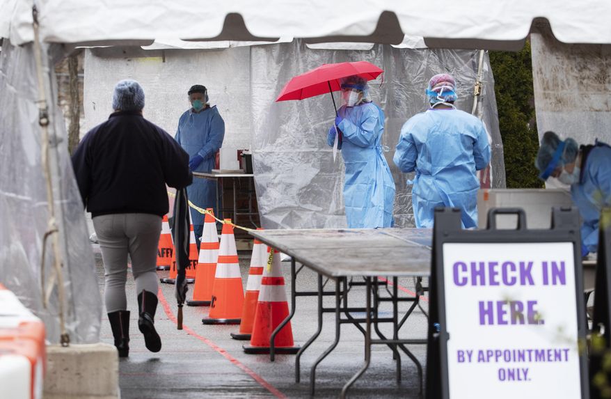 Dr. Beth Toolan, rear left, and nurse Natasha Viveiros, holding umbrella, wait for a patient at the state's first walk-up coronavirus testing site which opened Tuesday, April 21, 2020, in Providence, R.I. The site in the parking lot of the Robert L. Bailey IV Elementary School is an effort to bring testing to underserved communities, authorities say. The tests are by appointment only with a referral from a health care professional. (AP Photo/David Goldman)