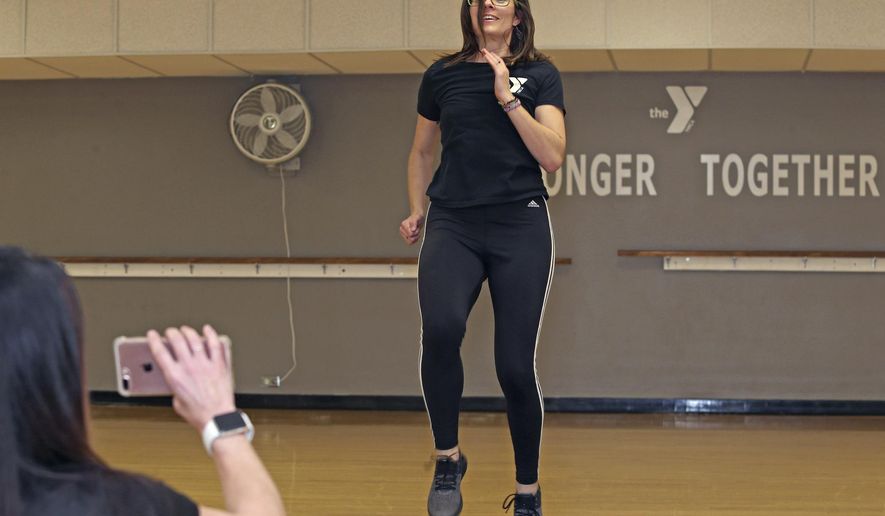 In this Wednesday, April 1, 2020 photo, trainer Heather Sommers, right, demonstrates a sample routine of the workouts she's been recording for Beth Hilbing, left, at the Quincy Family YMCA in Quincy, Ill. The Quincy Family YMCA has been posting actively posting videos on their Facebook page to keep members active while they're at home due to the shelter in place. (Jake Shane/Quincy Herald-Whig via AP)