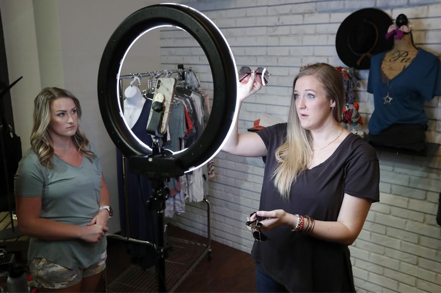 Assistant manager Kaila Yoachum, left, watches as product buyer Allison Scott promotes sunglasses during a live online video session in a corner of their store, Apricot Lane Boutique, at the Galleria in Dallas, Friday April 24, 2020. Scott said that online video sales was part of their business model, instituting the service to the clients several months before the COVID-19 pandemic. Scott said the online sales alone are what is helping their business survive through the health crisis. Lifting restrictions that were in place due to the new coronavirus pandemic, Gov. Greg Abbott announced that retail stores would be allowed to reopen and conduct curbside service beginning Friday. Galleria Dallas has begun a "Retail-To-Go" initiative which allows businesses at the retail location to re-open and conduct sales either online or over the phone. The process allows for retailers to deliver their product to their customers at a drive up lane that has been set up in front of the mall that is open daily from noon to 6 p.m. (AP Photo/Tony Gutierrez)