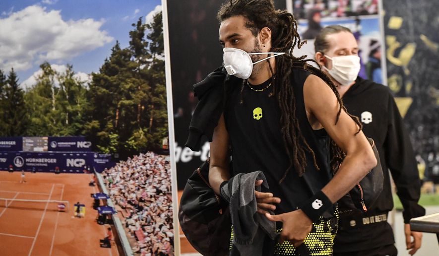 Germany's tennis player Dustin Brown wears a face mask as he walks by a poster at a pro-tennis tournament at a local base tennis academy in Hoehr-Grenzhausen, Western Germany, Friday, May 1, 2020. The professional tennis exhibition in the small village in the Westerwald is a rare exception to the global shutdown of sports during the coronavirus pandemic. Matches are played without line judges and without spectators, and broadcasted by remote cameras worldwide. (AP Photo/Martin Meissner)