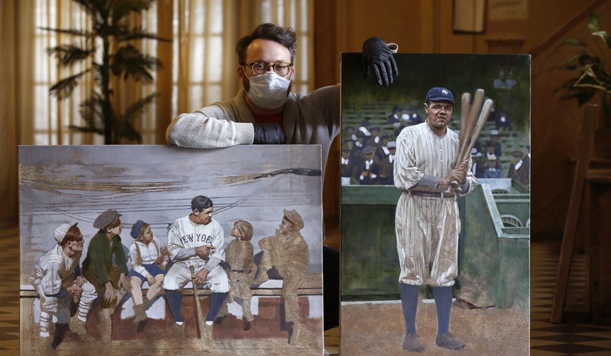 In this Wednesday, April 29, 2020, photo, Brooklyn-based baseball artist Graig Kreindler poses with two of his paintings of Babe Ruth, both unfinished, in the lobby of his residence in New York. Over 200 of Kreindler's paintings form the bulk of the exhibit "Black Baseball in Living Color:The Art of Graig Kreindler," at the Negro Leagues Baseball Museum in Kansas City, Mo. The museum is closed now, but expects to reopen early in June. Both paintings of Ruth are based on photographs. (AP Photo/Kathy Willens)