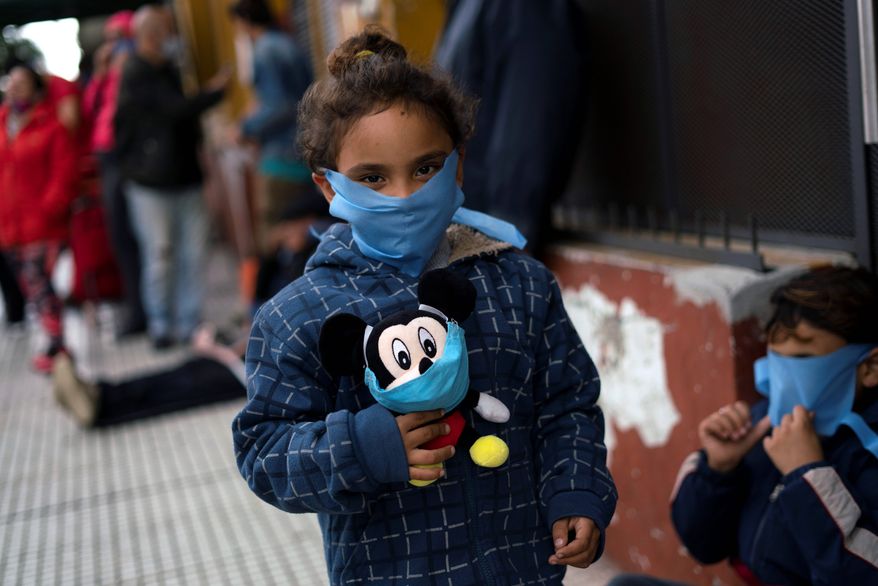 Wearing a protective face mask, a child poses for a photo holding her Mickey Mouse plush also fitted with a face mask, as she waits in line to receive free food from a group of neighbors who have formed to help people in need facing hardship due to the new coronavirus pandemic, in the financial district of Montevideo, Uruguay, Saturday, April 25, 2020. (AP Photo/Matilde Campodonico)