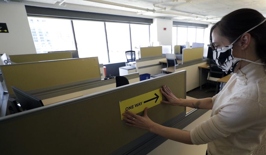 This Thursday, May 7, 2020, photo shows Interior Designer Stephanie Jones at the design firm Bergmeyer putting up a safe distancing reminder indicating one-way foot traffic to a cubicle at the firm's offices in Boston. When workers around the world eventually return to their desks, they’ll find myriad adjustments by their companies to reflect the post-pandemic “new normal” way of life, executives and experts say. (AP Photo/Steven Senne)
