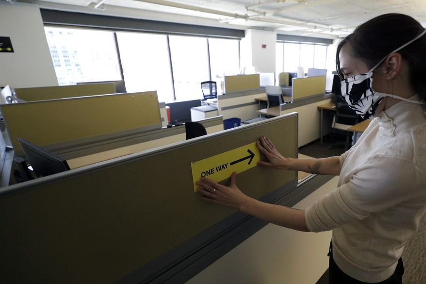 This Thursday, May 7, 2020, photo shows Interior Designer Stephanie Jones at the design firm Bergmeyer putting up a safe distancing reminder indicating one-way foot traffic to a cubicle at the firm's offices in Boston. When workers around the world eventually return to their desks, they’ll find myriad adjustments by their companies to reflect the post-pandemic “new normal” way of life, executives and experts say. (AP Photo/Steven Senne)