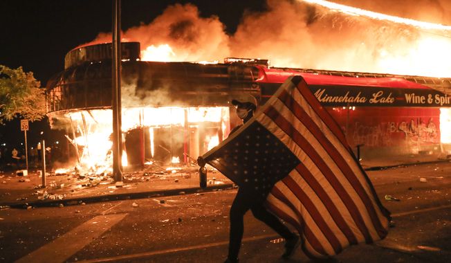 A protester carries a U.S. flag upside, a sign of distress, next to a burning building, early Friday, May 29, 2020, in Minneapolis. Protests over the death of George Floyd, a black man who died in police custody Monday, broke out in Minneapolis for a third straight night. (AP Photo/Julio Cortez)
