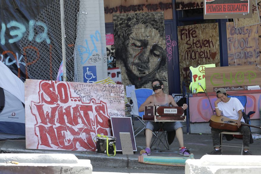 Two people perform music next to a sign in front of the Seattle Police Department's East Precinct building in the CHOP (Capitol Hill Occupied Protest) zone reads "So What's Next?" Thursday, June 25, 2020, in Seattle. The area has been occupied by Black Lives Matter protesters since police pulled back from the building following violent clashes with demonstrators, and the future of the protest area has been in question since Seattle Mayor Jenny Durkan said earlier in the week that police intend to eventually return to the building. (AP Photo/Ted S. Warren)