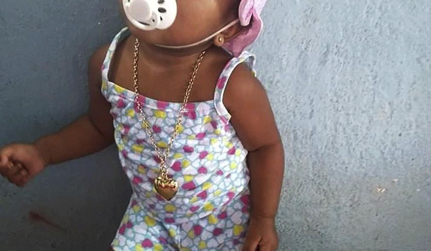In this Feb. 10, 2020, photo, provided by Andréa de Sousa, her daughter Vitoria Gabrielle looks up while leaning against a wall, in Rio de Janeiro, Brazil. The girl with a constant smile celebrated her first birthday in February, but after recovering from viral meningitis, Vitoria Gabrielle suffered gastrointestinal problems that sent her from her mother&#39;s barely furnished hilltop home back to the hospital several times for treatment. It was during an April hospital stay that de Sousa suspects her daughter was infected with the coronavirus that was just starting to circulate in Rio and Brazil. Vitoria Gabrielle died in June. (Courtesy of Andréa de Sousa via AP)