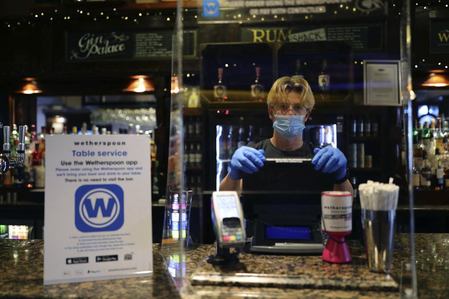 A member of staff wearing PPE, stands behind a screen as he waits to serve drinkers at the reopening The Toll Gate, a Wetherspoons pub in Hornsey, north London, as coronavirus lockdown restrictions are eased across the country, Saturday July 4, 2020. Restrictions which were imposed on March 23 have been eased allowing  businesses including pubs, restaurants and hair salons, to reopen to members of the public with measures in place to prevent the spread of the coronavirus. (Aaron Chown/PA via AP)