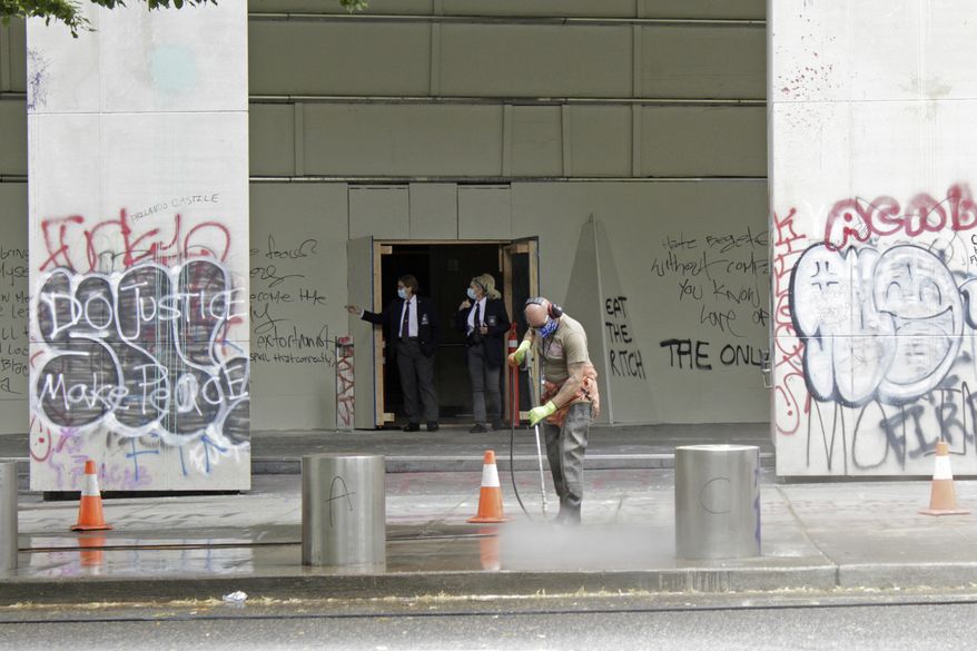 In this July 8, 2020, file photo, a worker washes graffiti off the sidewalk in front of the Mark O. Hatfield Federal Courthouse in downtown Portland, Ore., as two agents with the U.S. Marshals Service emerge from the boarded-up main entrance to examine the damage. Oregon's largest city is in crisis as violent protests have wracked downtown for weeks. (AP Photo/Gillian Flaccus, File) ** FILE **