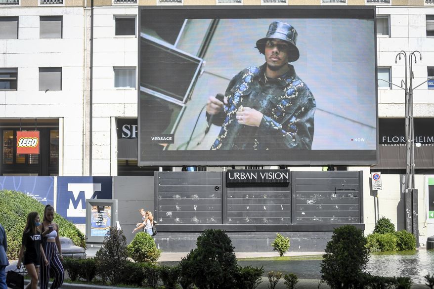 Pedestrians pass by a screen showing a Versace model, during the Milan Digital Fashion Week, in Milan, Italy, Thursday, July 16, 2020. Forty fashion houses are presenting previews of menswear looks for next spring and summer and pre-collections for women in digital formats, due to concerns generated by the COVID-19. (Claudio Furlan/LaPresse via AP)