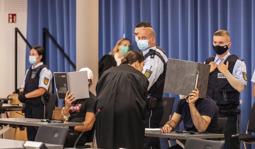 Two defendants are holding folders in front of their faces as a screen, while a defense attorney is talking to the right defendant and judicial officers are standing behind the defendants at a trial in Freiburg, Germany, Thursday, July 23, 2020. Most of the eleven defendants have been sentenced to prison for the gang rape of an 18-year-old girl in Freiburg in mid-October 2018. In order to comply with the Corona rules, the trial did not take place in a court, but in a concert hall. (Philipp von Ditfurth/dpa via AP)