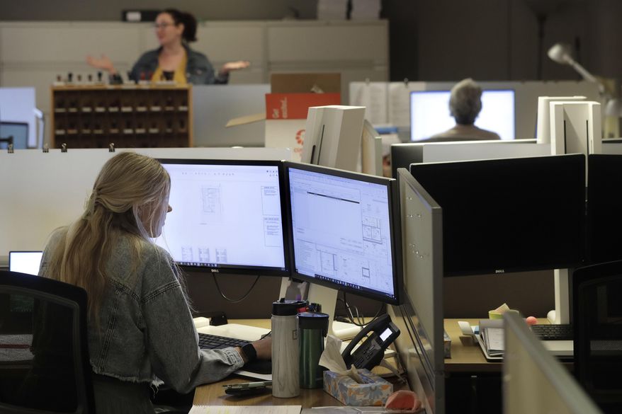 Architectural designer Erica Shannon, front, works at a computer as accounting manager Andrea Clark, top, speaks with a colleague at the design firm Bergmeyer, Wednesday, July 29, 2020, at the company's offices, in Boston. Around the U.S,. office workers sent home when the coronavirus took hold in March are returning to the world of cubicles and conference rooms and facing certain adjustments: masks, staggered shifts, limits on how many people can be there at any one time, spaced-apart desks, daily questions about their health, closed break rooms, sanitizer everywhere. (AP Photo/Steven Senne)