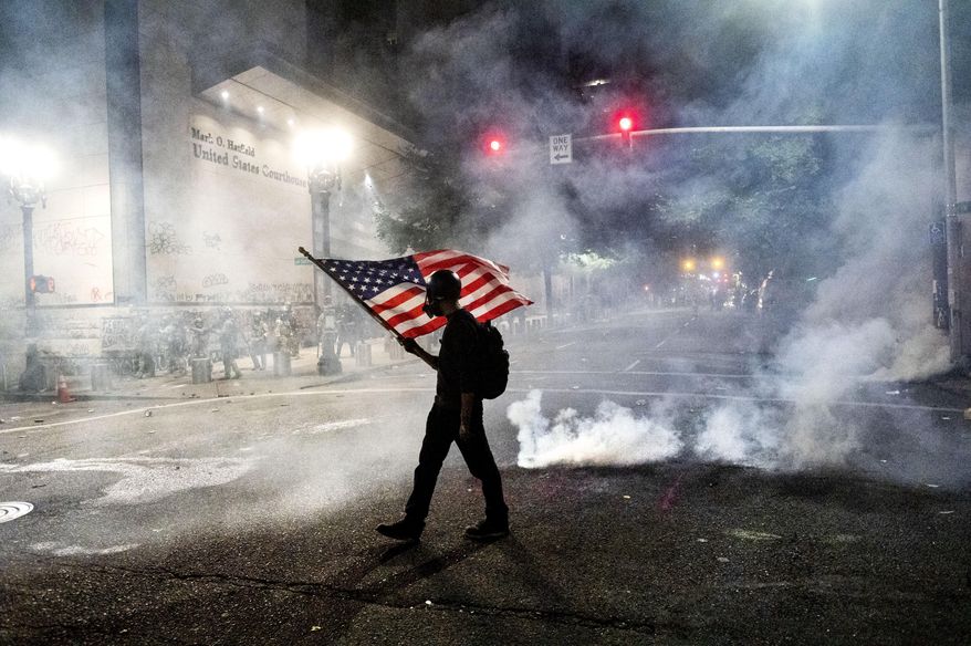 In this July 21, 2020, file photo, a Black Lives Matter protester carries an American flag as teargas fills the air outside the Mark O. Hatfield United States Courthouse in Portland, Ore. (AP Photo/Noah Berger, File)