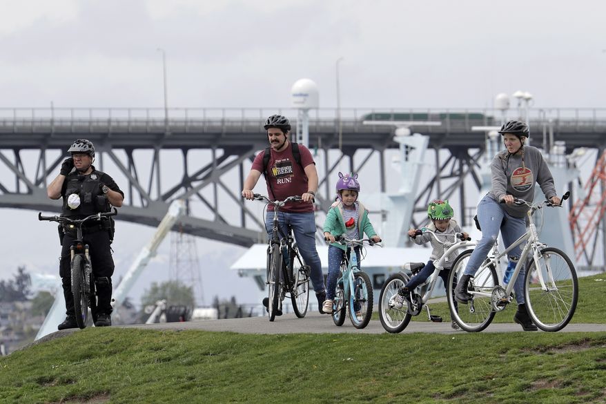 A Seattle Police Officer on a bicycle watches from a hill as people ride by at Gas Works Park in Seattle, Sunday, April 26, 2020. The park and others in Seattle were open Sunday, but officials sought to prevent the spread of the coronavirus by urging people to keep moving and not to gather in large groups. (AP Photo/Ted S. Warren)
