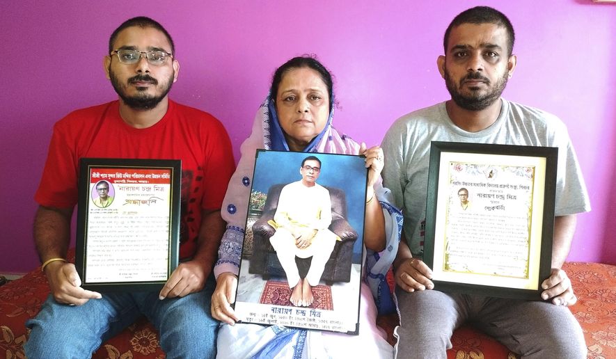 Anindita Mitra, 61, flanked by her sons Satyajit Mitra, right and Abhijit Mitra, pose with portraits of her husband late Narayan Mitra, at her house in Silchar, India, Sunday, Sept. 13, 2020. Narayan Mitra, wasn&#39;t listed among those killed by the coronavirus that authorities put out daily because the test results confirming COVID-19 arrived after his death. In India, people who die with other preexisting conditions are often not counted as COVID-19 deaths, while only those who test positive for the virus before dying are included in the official tally in many states. (AP Photo/Joy Roy)