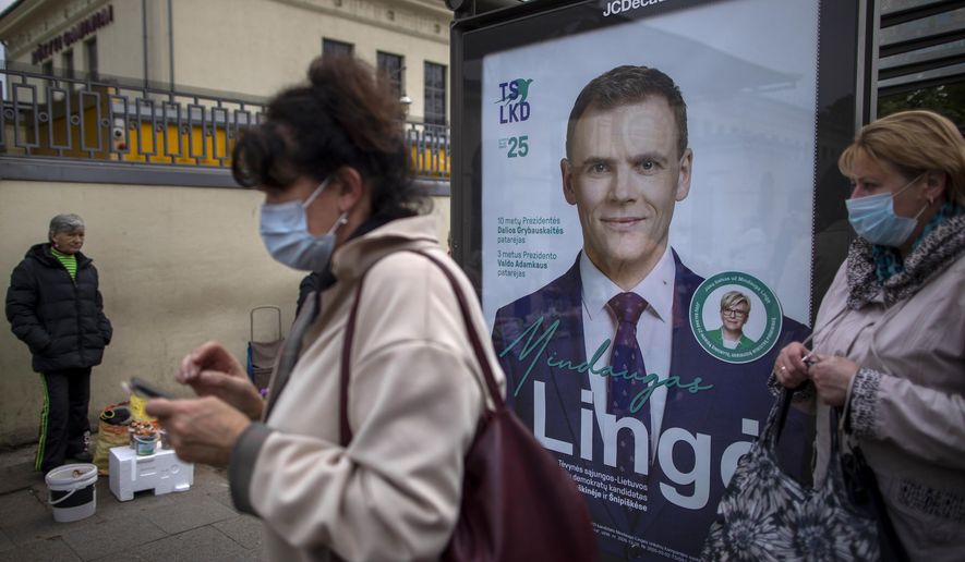 Women wearing face masks to protect against coronavirus wait at a bus stop, with a poster displaying Lithuania's Homeland Union – Christian Democrats (TS-LKD) party member Mindaugas Linge, in Vilnius, Lithuania, Thursday, Oct. 8, 2020. Lithuanians are voting in the first round of a parliamentary election on Sunday. (AP Photo/Mindaugas Kulbis)