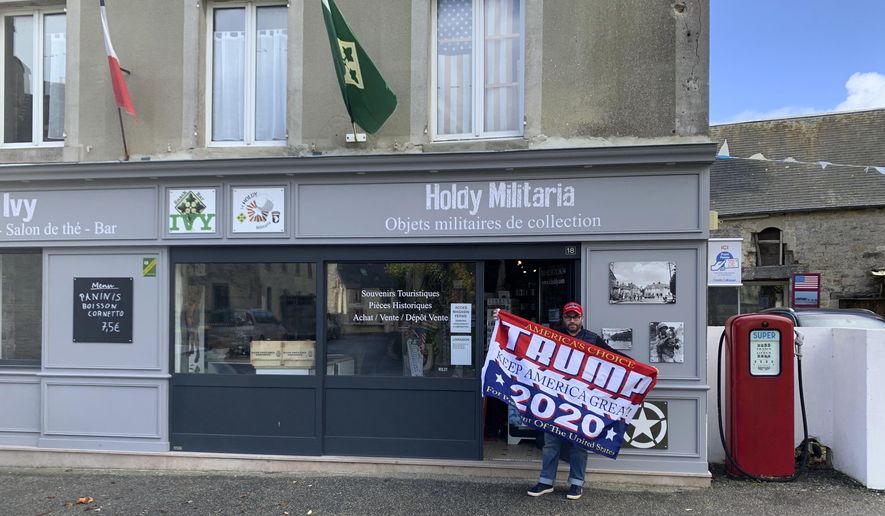Philippe Tanne, of France, holds a Trump 2020 flag outside the military memorabilia store he runs in the Normandy town of Sainte-Marie-du-Mont, one of the sites of D-Day invasion in 1944, Tuesday, Nov. 3, 2020. Tanne, a French former soldier who hopes Trump wins reelection, is among the multitudes of people across the globe for whom the U.S. election is not a far-away happening in a far-away land but an impossible-to-ignore big deal for the planet. (Courtesy of Philippe Tanne via AP)