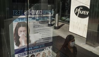 An ad for COVID-19 testing reflects on glass at a bus stop, as pedestrians walk past Pfizer world headquarters in New York on Monday Nov. 9, 2020. Pfizer says an early peek at its vaccine data suggests the shots may be 90% effective at preventing COVID-19, but it doesn&#x27;t mean a vaccine is imminent. (AP Photo/Bebeto Matthews)