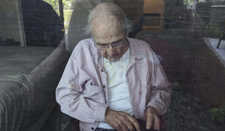 This June 7, 2020, photo provided by June Linnertz shows her father, James Gill, seen through a window at Cherrywood Pointe nursing home in Plymouth, Minn. As more than 90,000 of America’s long-term care residents have died in the coronavirus pandemic, advocates for the elderly say a tandem wave of death separate from the virus has quietly claimed tens of thousands more, often because overburdened workers haven’t been able to give them the care they need. Gill died of Lewy Body Dementia, according to a copy of his death certificate provided to the AP. Linnertz always expected her father to die of the condition, which causes a progressive loss of memory and movement, but never thought he would end his days in so much needless pain. (June Linnertz via AP)