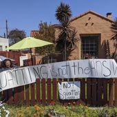 In this March 30, 2020, file photo, resident Marie Salas, left, sets a banner on her home fence reading &amp;quot;Squatting is not the Answer,&amp;quot; across the street of a formerly vacant home that was recently taken by a group of homeless mothers in the El Sereno neighborhood of Los Angeles. A new group of homeless people who had taken over several empty, state-owned houses in Los Angeles was forcefully removed by California Highway Patrol officers hours later amid protests by dozens of community activists. (AP Photo/Damian Dovarganes, File)