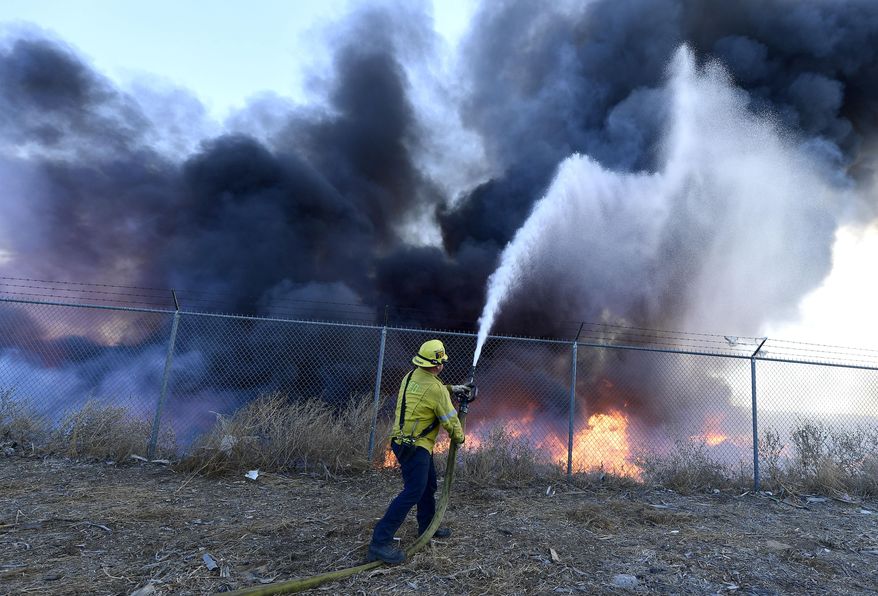 A firefighter battles a mulch and pallet fire burning out of control, fanned by Santa Ana winds in and around a recycling yard near Wilson Street and Fleetwood Drive in Riverside, Calif., Thursday, Dec. 3, 2020. Firefighters from both Riverside and San Bernardino County, along with assistance from Colton, Rialto and Riverside City Fire fought the blaze. (Will Lester/The Orange County Register via AP)