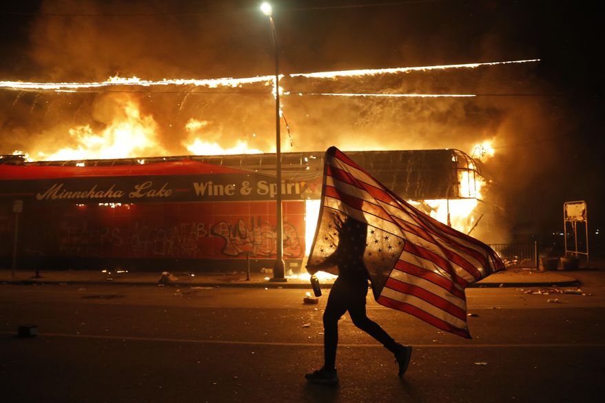 A protester carries a U.S. flag upside down as he walks past a burning building in Minneapolis on May 28, 2020, during a protest over the death of George Floyd, a Black man who died after a white Minneapolis police officer pressed a knee into his neck for several minutes. (AP Photo/Julio Cortez)