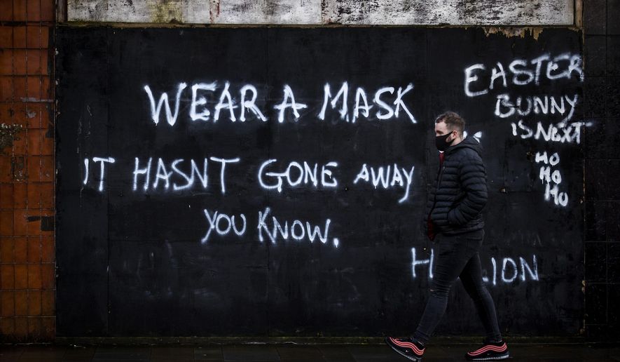 A man wearing a face covering walks past graffiti on the Lower Newtownards Road in Belfast with a message reading &#39;Wear a mask, it hasn&#39;t gone away you know&#39; Friday, Jan. 1, 2021. (Liam McBurney/PA via AP)