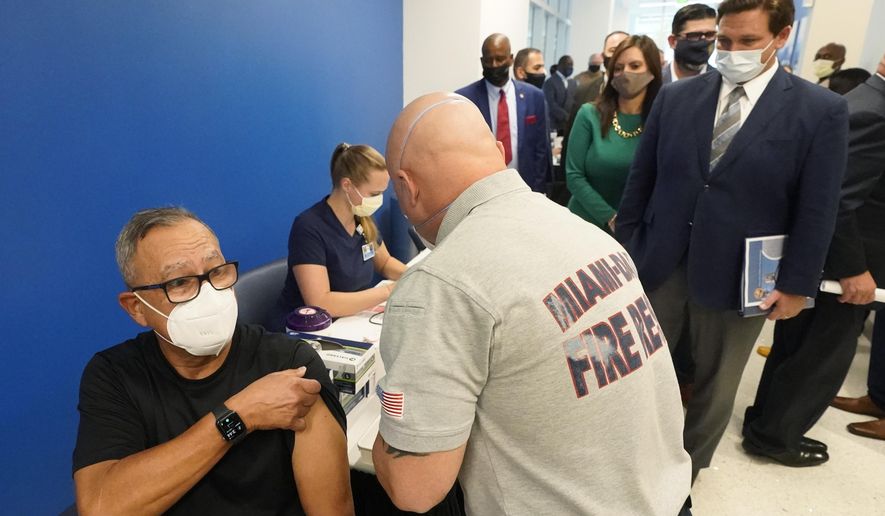 FILE - In this Jan. 4, 2021, file photo, Florida Gov. Ron DeSantis, right rear, watches as Carlos Dennis, left, 65, rolls up his sleeve so that Miami-Dade County Fire Rescue paramedic, Capt. Javier Crespo, can administer a COVID-19 vaccine shot at Jackson Memorial Hospital in Miami. Florida was one of the first states to throw open vaccine eligibility to members of the general public over 65, leading to rumors that tourists and day-trippers are swooping into the state solely for the jab.  (AP Photo/Wilfredo Lee, File)