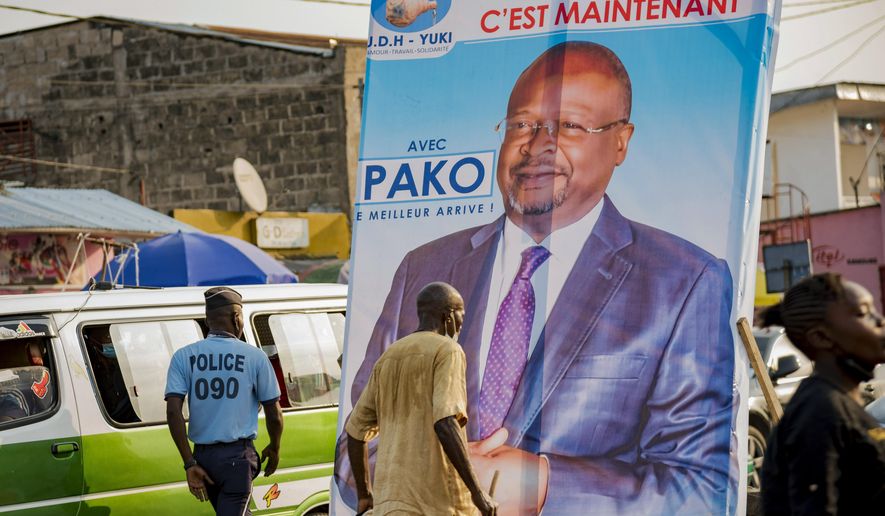 FILE- In this Sunday March 7, 2021 file photo, people walk past an election poster featuring opposition presidential candidate Guy Brice Parfait Kolelas, in downtown Brazzaville, Congo. Kolelas, who was hospitalized with COVID-19 complications on election day, has died, a spokesman said Monday, March 22, 2021. The 61-year-old politician was last seen in a video circulating Saturday on social media in which he told supporters he was “fighting death.” Aides later said he was been flown to France for further treatment. Spokesman Justin Nzoloufoua confirmed his death Monday to The Associated Press. (AP Photo/Lebon Chansard Ziavoula, File)