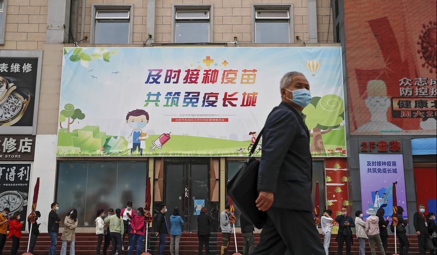 A man wearing a face mask to help curb the spread of the coronavirus walks by masked residents lining up for COVID-19 vaccine at a vaccination site with a board displaying the slogan, "Timely vaccination to build the Great Wall of Immunity together" in Beijing, Wednesday, April 21, 2021. (AP Photo/Andy Wong)