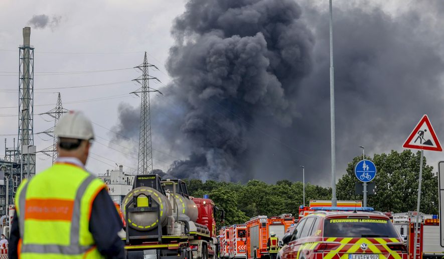 Emergency vehicles of the fire brigade, rescue services and police stand not far from an access road to the Chempark over which a dark cloud of smoke is rising in Leverkusen, Germany, Tuesday, July 27, 2021. After an explosion, fire brigade, rescue services and police are currently in a large-scale operation, the police explained. (Oliver Berg/dpa via AP)