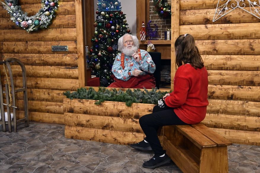 Santa, Sid Fletcher, sits behind a glass barrier as he hears Kendra Alexander of St. James, Minn., during her visit Monday, Nov. 15, 2021, at The Santa Experience at the Mall of America in Bloomington, Minn. Where allowed, some malls and big-box stores are offering Santa's guests a choice of full contact or social distance. A microphone in the green plays picks up conversations. (AP Photo/Jim Mone)