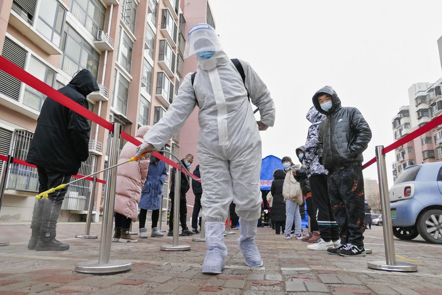 A worker wearing a protection suit sprays disinfectant as residents wearing face masks to protect from the coronavirus line up for the coronavirus test during a mass testing in north China's Tianjin municipality, Sunday, Jan. 9, 2022. Tianjin, a major Chinese port city near the capital Beijing, began mass testing of its 14 million residents on Sunday, after a cluster of a dozen of children and adults tested positive for COVID-19, including a few with the omicron variant. (Chinatopix Via AP)