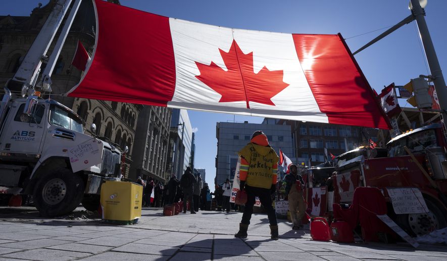 A protester stands under a giant Canadian flag, Monday, Feb. 14, 2022 in Ottawa. The protesters are decrying federal vaccine mandates and provincial COVID-19 restrictions aimed at slowing the spread of the virus, some of which are being rolled back by provinces. (Adrian Wyld/The Canadian Press via AP)