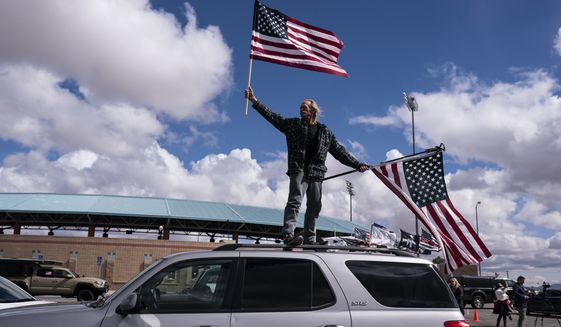 Jeff Cassil waves two American flags from the roof of his car at the beginning of a trucker caravan to Washington, D.C., called The People&#39;s Convoy Wednesday, Feb. 23, 2022, in Adelanto, Calif. A small convoy of truckers demanding an end to coronavirus mandates began a cross-country drive from California to the Washington, D.C., area on Wednesday. (AP Photo/Nathan Howard)