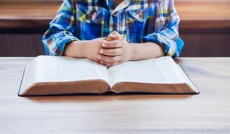 A child praying on an open Bible at a wooden table. (Photo credit: Freedom Studio via Shutterstock)