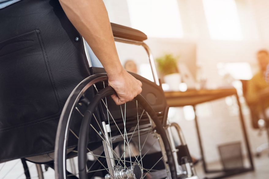 A disabled man is sitting in a wheelchair. He holds his hands on the wheel. Nearby are his colleagues. File photo credit: VGstockstudio via Shutterstock