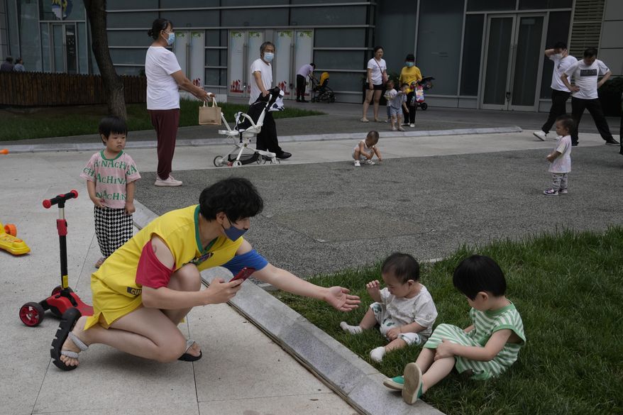 A woman wearing a mask tries to take photos for children as they play on the lawn, Wednesday, July 6, 2022, in Beijing. (AP Photo/Ng Han Guan)